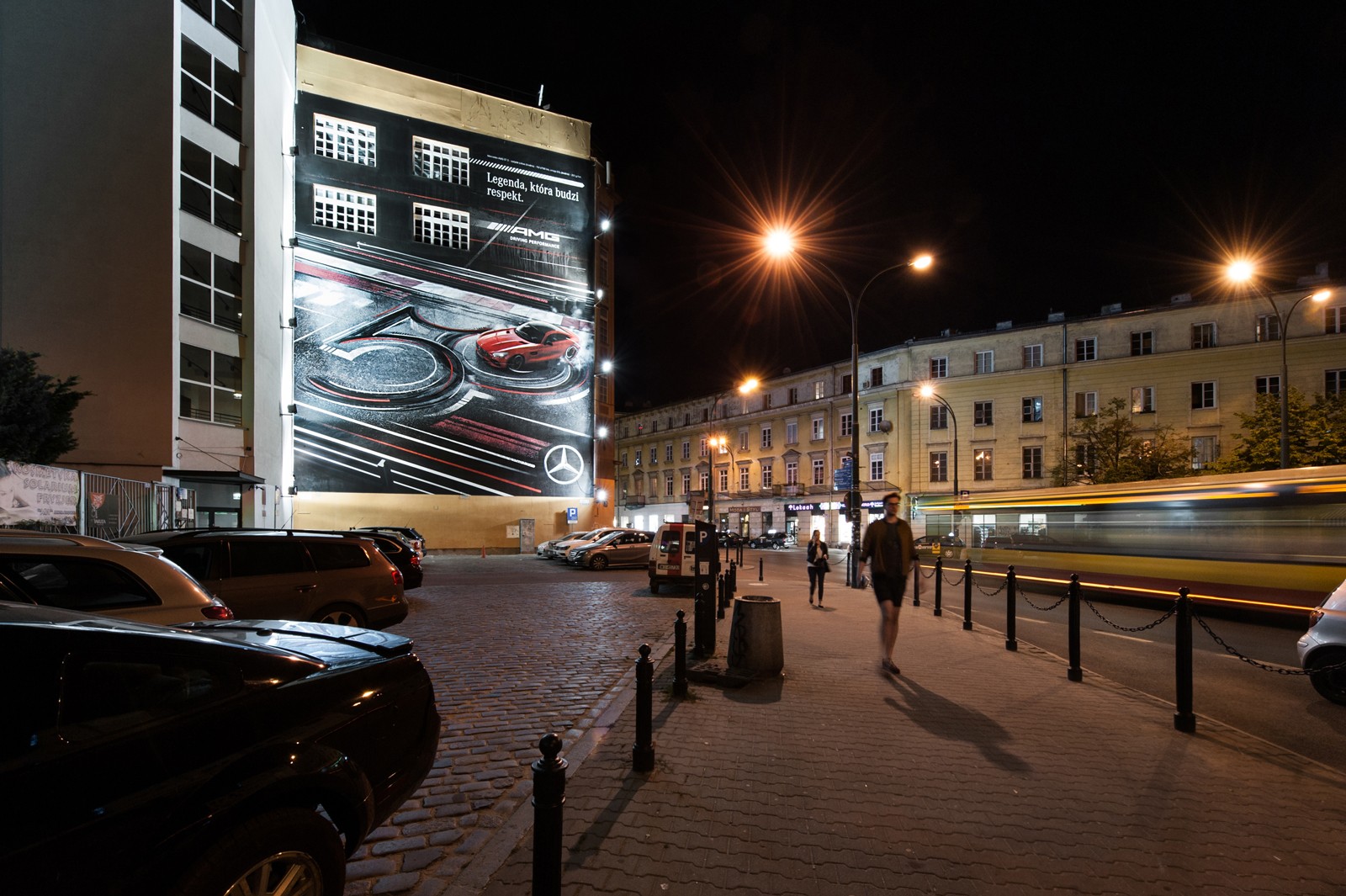Widok nocny z ulicy Kruczej na Dom Towarowy Braci Jabłkowskich przy ulicy Brackiej 25 w Warszawie z namalowanym muralem dla Mercedes Benz Polska | Kampania z wykorzystaniem murali na zlecenie Mercedes-Benz Polska | Portfolio
